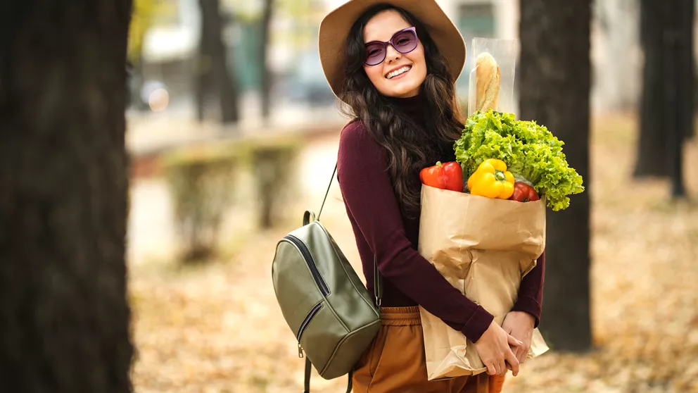 Mujer sonriente con sombrero y gafas de sol llevando una bolsa de productos